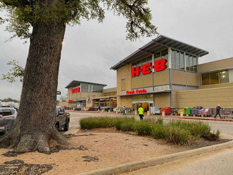A large H-E-B grocery store with red signage, a tree in the foreground, and people walking near the entrance on a cloudy day. Pallets and shopping carts are near the store’s front.