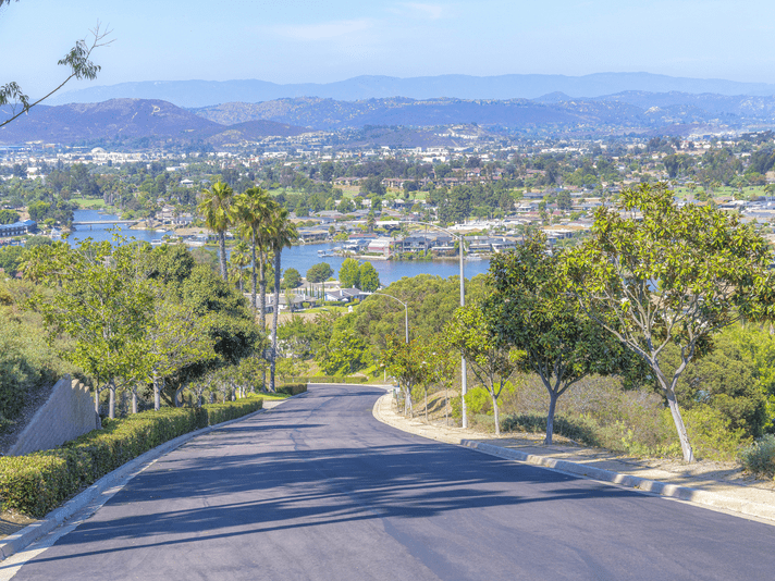 A paved road curves downhill, lined with trees and greenery, leading to a scenic view of a lake, houses, and rolling hills under a clear blue sky.