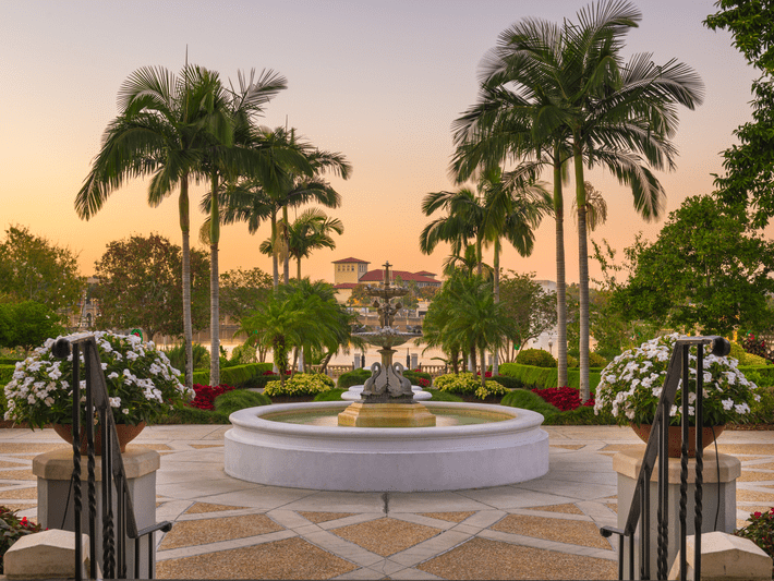 A peaceful garden at sunset with a circular fountain in the center, surrounded by palm trees, colorful flowers, greenery, and a view of water and buildings in the background.