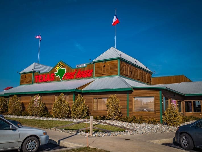 A Texas Roadhouse restaurant with a wooden exterior, silver metal roof, and two flags on top—one American and one Texas. The logo features a green outline of Texas with a yellow star and cowboy hat. Cars are parked in front.