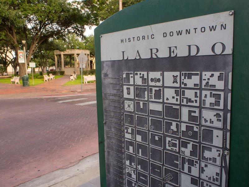 A metal sign displays a map of historic downtown Laredo, Texas, with a park, benches, and trees in the background on a sunny day.