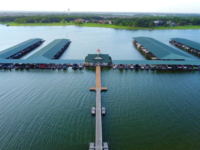 Aerial view of a marina with covered boat docks on a lake, a central pavilion at the end of a long pier, and green shoreline with houses and trees in the background.
