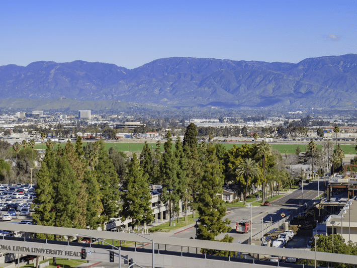 A cityscape with green fields, trees, parking lots, and buildings in the foreground, set against a backdrop of tall mountains under a clear blue sky.