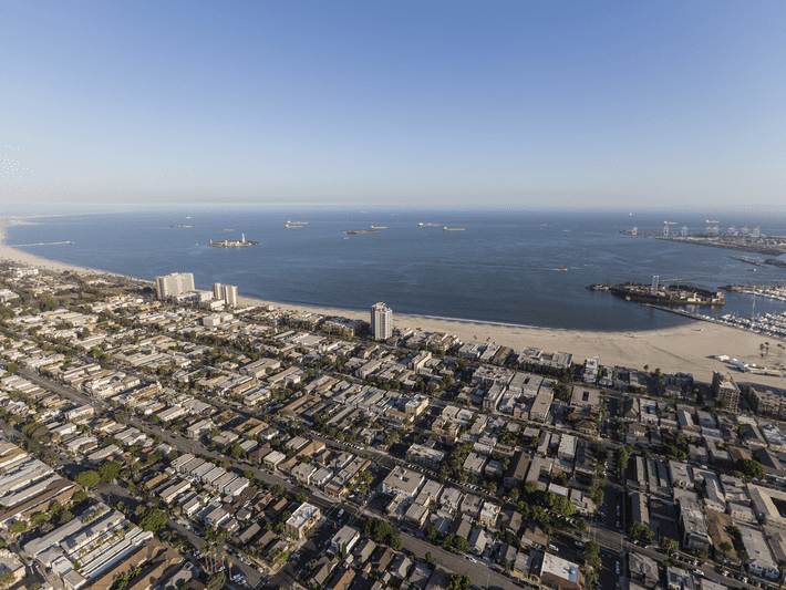 Aerial view of a coastal city with rows of buildings, sandy beach, calm ocean, and several ships anchored offshore under a clear blue sky.
