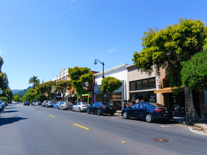 A sunny day view of a small town street lined with parked cars, trees, and storefronts. The sky is clear and blue, and a palm tree is visible in the distance.
