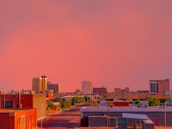 A cityscape at sunset with a pink and orange sky, low-rise buildings in the foreground, and taller buildings in the background. The streets are quiet and bathed in warm, colorful light.