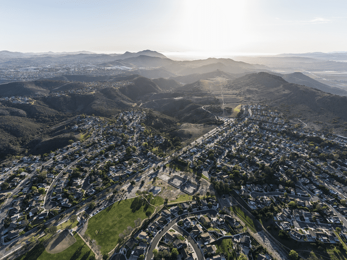 Aerial view of a suburban neighborhood with winding roads, houses, green parks, and hills in the background under a clear sky with the sun low on the horizon.