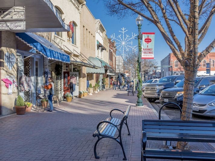 A sunny street scene in a small downtown area with shops, parked cars, benches, leafless trees, people walking, and a red banner on a lamppost that says CHRISTMAS beside a snowflake decoration.