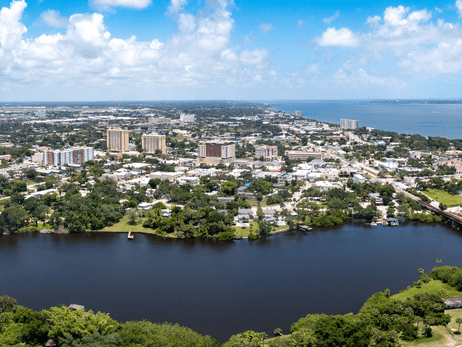 Aerial view of a coastal city with tall buildings, green trees, and a wide river in the foreground, leading out to a blue ocean under a partly cloudy sky.