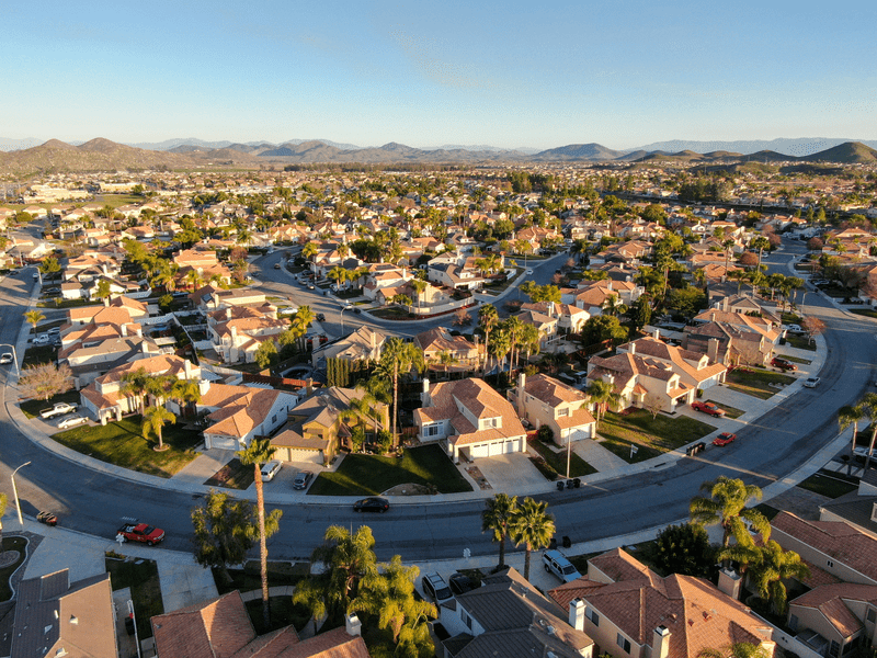 Aerial view of a suburban neighborhood with rows of houses, tidy lawns, curved streets, and scattered palm trees, set against distant mountains under a clear sky.