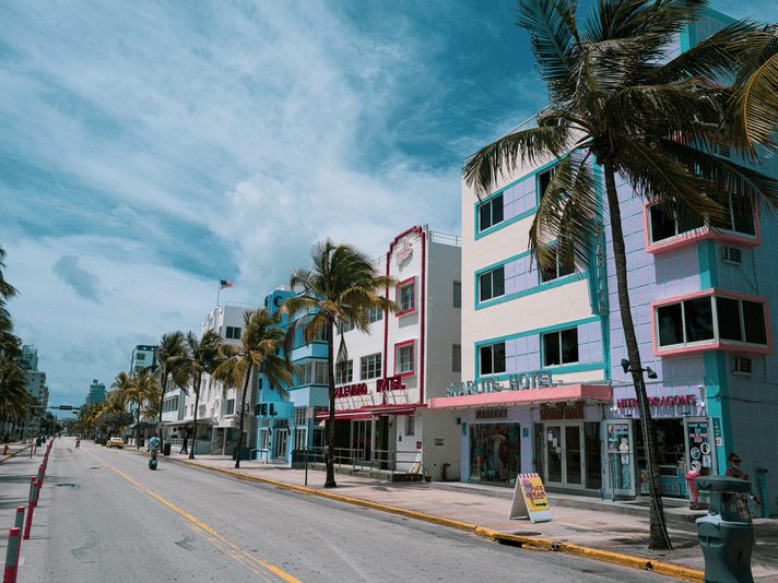 A nearly empty street in Miami Beach, Florida, lined with art deco hotels and palm trees under a partly cloudy blue sky. One person rides a scooter on the road.