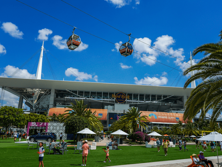 People walk and relax on a grassy area in front of Hard Rock Stadium, with palm trees and white tents nearby, as gondola cable cars move overhead against a blue sky with scattered clouds.
