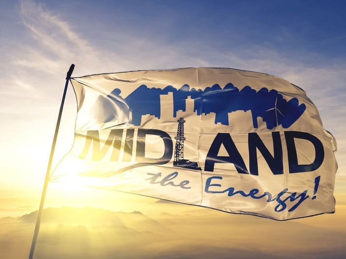 A white flag with blue graphics of a city skyline, oil rig, and wind turbine reads MIDLAND the Energy! as it waves in the wind against a sunrise or sunset sky.