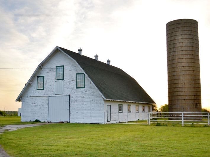 A white barn with a green roof stands beside a tall, cylindrical silo on a grassy field, with a white fence and a bright sky in the background.