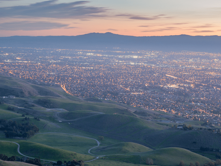A scenic view of rolling green hills in the foreground with a cityscape full of lights spread out in the valley below at dusk, with mountains visible in the background under a colorful sky.