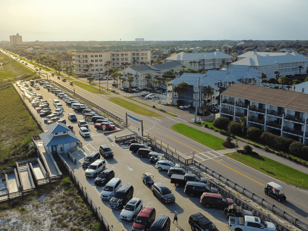 Aerial view of a crowded parking lot near a beach, with rows of parked cars, adjacent buildings, and a road running parallel to the shoreline under a clear sky.