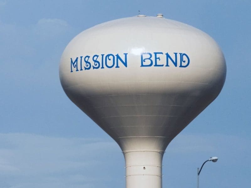 A large white water tower with the words MISSION BEND painted in blue near the top, set against a partly cloudy blue sky.