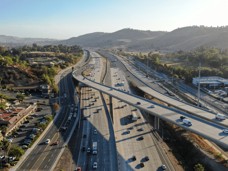 Aerial view of a wide freeway with multiple lanes and overpasses, light to moderate traffic, surrounded by hills, trees, and some buildings on a clear day.