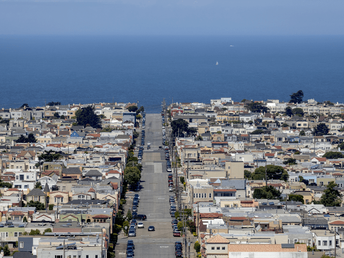 A wide street lined with houses stretches toward the blue ocean under a clear sky, with rows of buildings on both sides and a few cars parked along the road.