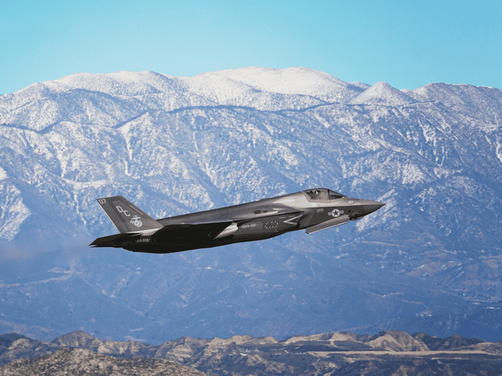 A military fighter jet flies above mountainous terrain, with snow-capped peaks and a clear blue sky in the background.