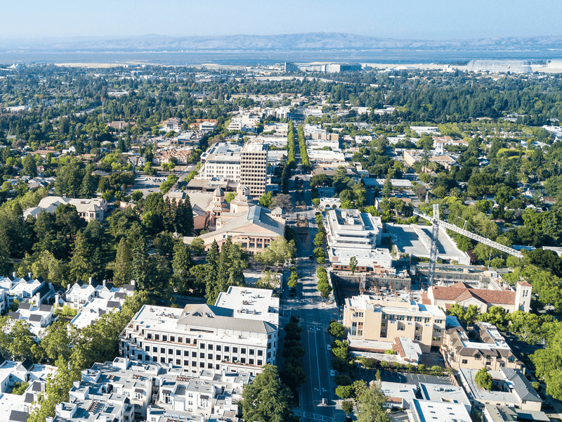 Aerial view of a city with tree-lined streets, various buildings, and distant mountains under a clear blue sky.