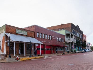 A row of historic brick buildings with shops, including a mercantile store, lines a quiet, brick-paved street on an overcast day. A few parked cars are visible along the street.