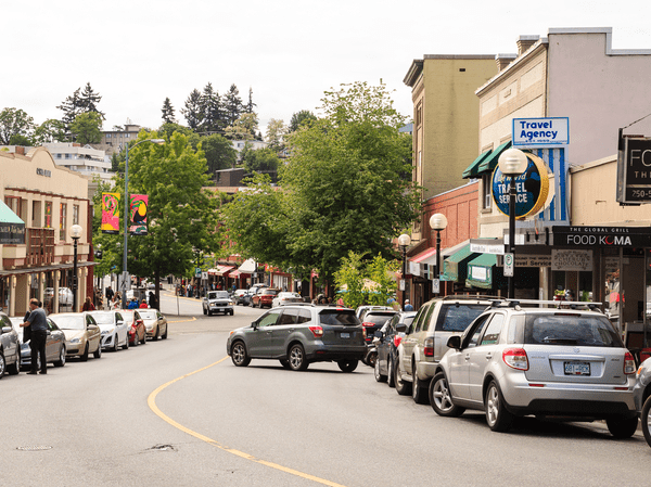 A curved street in a small downtown area lined with parked cars, storefronts, and trees. People walk on the sidewalks, and a car is turning into a parking spot. Signs for cafés and a travel agency are visible.