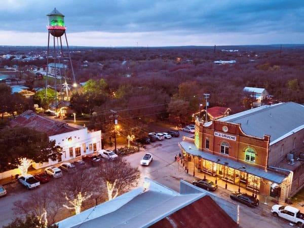 Aerial view of a small town at dusk, featuring a lit-up water tower, historic brick buildings, street lights, parked cars, and trees, with holiday lights decorating the area.