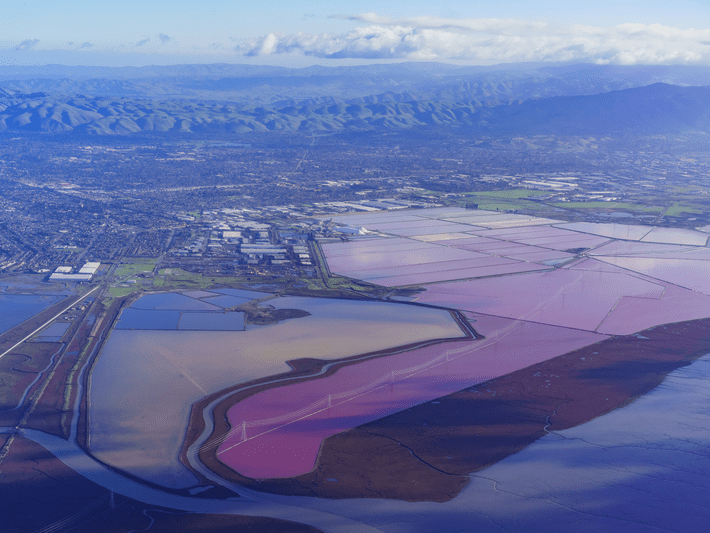 Aerial view of salt ponds near a coastline, featuring rectangular pools in shades of pink, brown, and blue, with a city, green fields, and mountains in the background under a partly cloudy sky.
