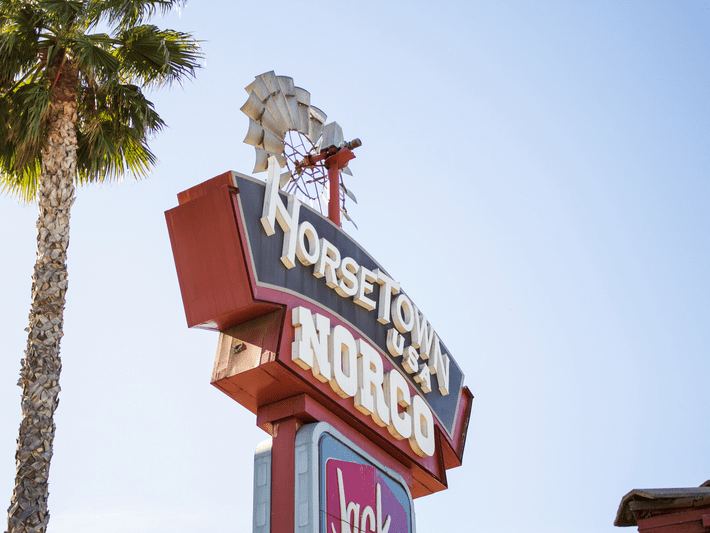 A vintage roadside sign with a windmill on top reads Horse Town U.S.A. Norco against a clear blue sky. A palm tree is on the left, and part of a fast food sign is visible at the bottom.