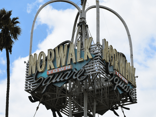 A vintage metal sign reads Norwalk Town Square in large letters, mounted on a tall structure against a partly cloudy sky with a palm tree visible to the left.