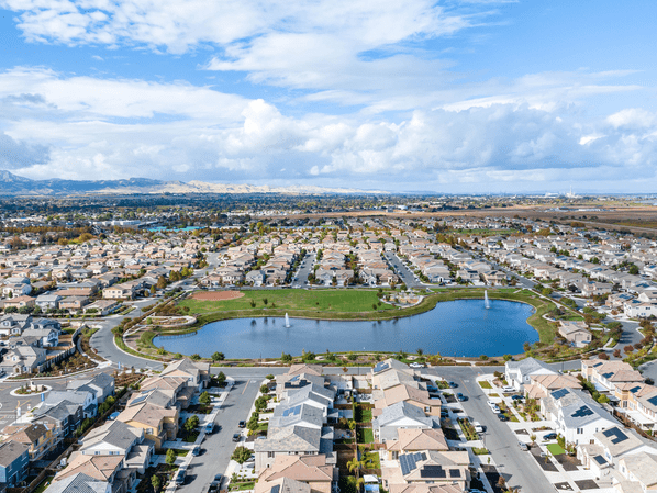 Aerial view of a suburban neighborhood with rows of houses surrounding a large pond and green park area, under a partly cloudy sky.