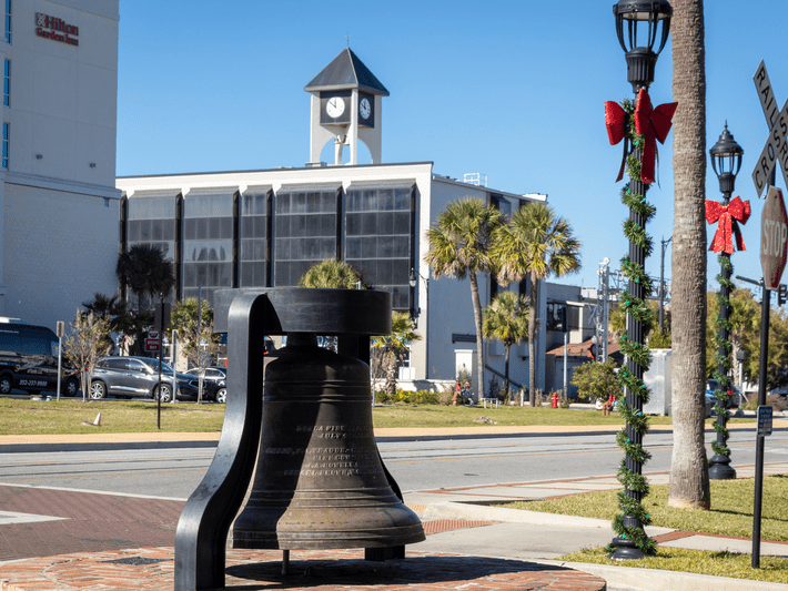 A large bronze bell on display in a plaza with a clock tower in the background; nearby streetlamps are decorated with green garlands and red bows, and palm trees line the street under a clear blue sky.