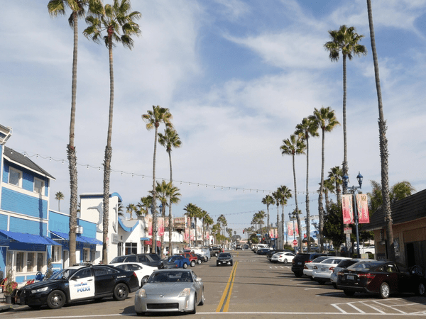 A sunny street lined with tall palm trees, parked cars, and colorful buildings, including a blue one on the left. A silver car drives along the road under a partly cloudy sky.