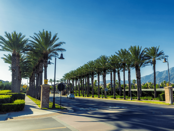 A sunny street lined with tall palm trees and streetlights, with mountains visible in the background and a single car driving on the road. The sky is clear and blue.