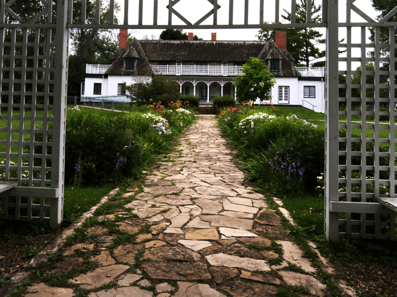 A stone pathway leads through lush gardens to a large white historic house with a porch and gabled roof, framed by a wooden trellis in the foreground.