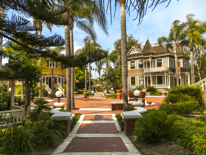 A brick walkway lined with palm trees leads to Victorian-style houses and lush green landscaping under a clear blue sky. White globe lamps and manicured bushes accent the picturesque, peaceful neighborhood.
