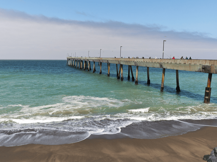 A long pier extends over the turquoise ocean, with waves gently crashing onto the sandy beach under a partly cloudy sky. People can be seen walking and fishing along the pier.