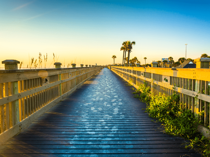 A wooden boardwalk lined with railings stretches toward the horizon, flanked by green plants and palm trees under a bright, clear blue sky at sunrise or sunset.