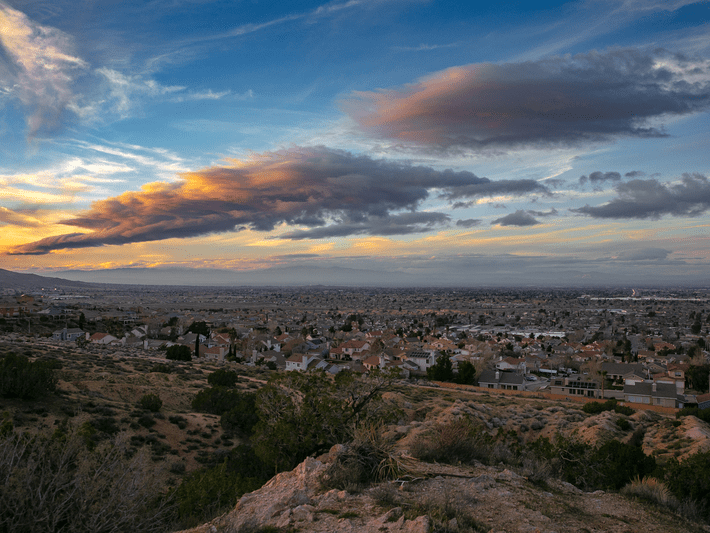 A wide view of a suburban neighborhood with houses spread out across a flat landscape under a dramatic sky with streaks of clouds lit by the setting sun. Bushes and rocky terrain appear in the foreground.
