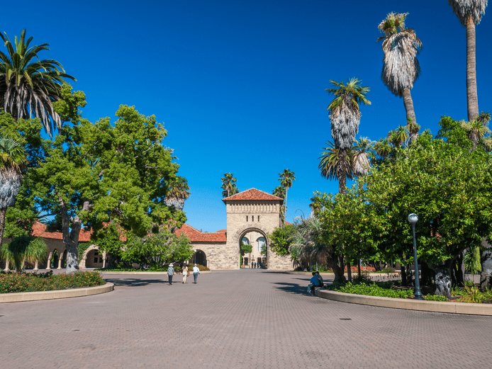 People walk near a stone archway and red-roofed buildings surrounded by palm trees and greenery under a clear blue sky on a sunny day.