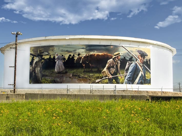 A large cylindrical white building displays a mural of pioneers and cattle crossing a muddy field, with a woman in a bonnet and men in hats leading the way. Yellow wildflowers grow in the grassy area in front.