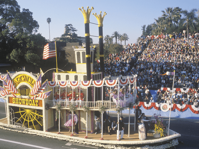 A parade float decorated as a steamboat with “Arcadia Showboat” sign, American flags, and people in colorful costumes passes by grandstands filled with spectators. Trees are visible in the background.