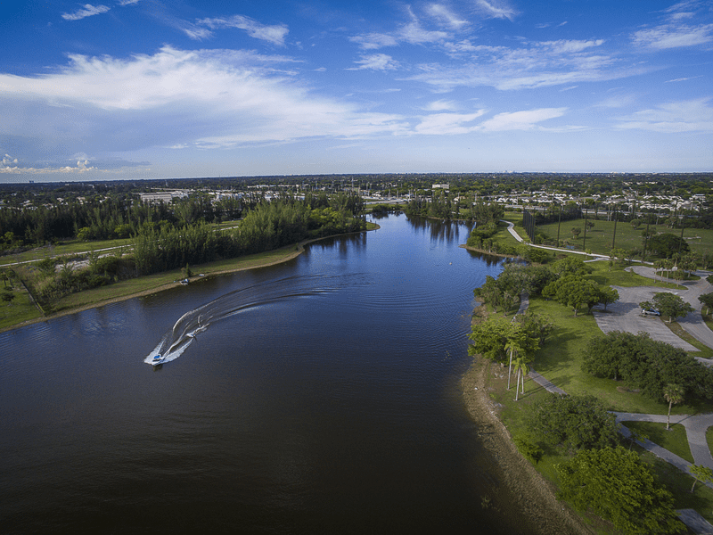 Aerial view of a river with a speedboat creating a wake, surrounded by green trees and grassy areas under a partly cloudy blue sky, with a town in the distance.