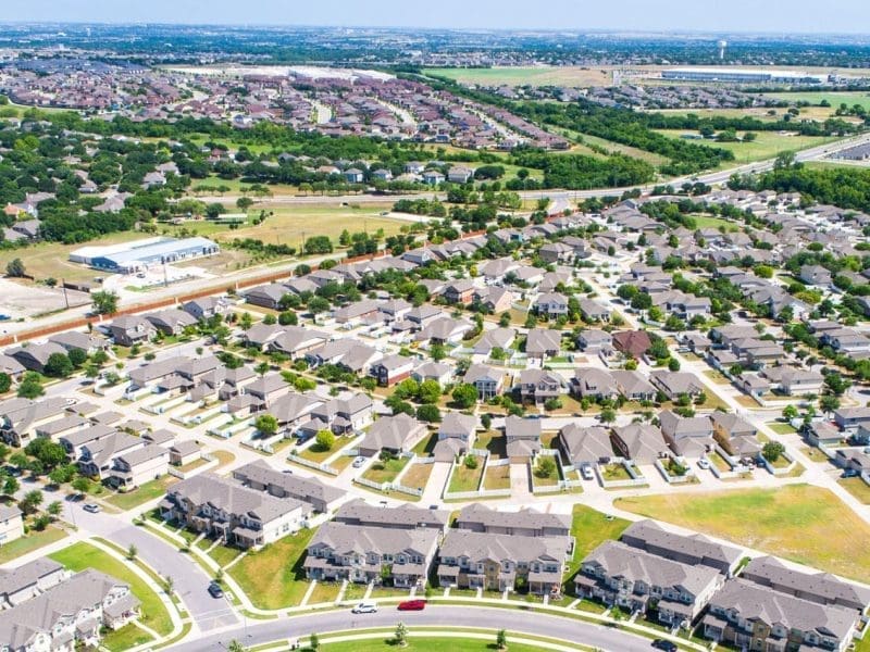 Aerial view of a suburban neighborhood with rows of houses, green lawns, winding streets, and trees. More housing developments and open land are visible in the background under a clear sky.