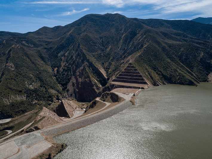 Aerial view of a dam and reservoir set against steep, green mountains under a partly cloudy sky. The dam’s structure and terraced slopes are visible, with the reservoir’s water spanning the foreground.
