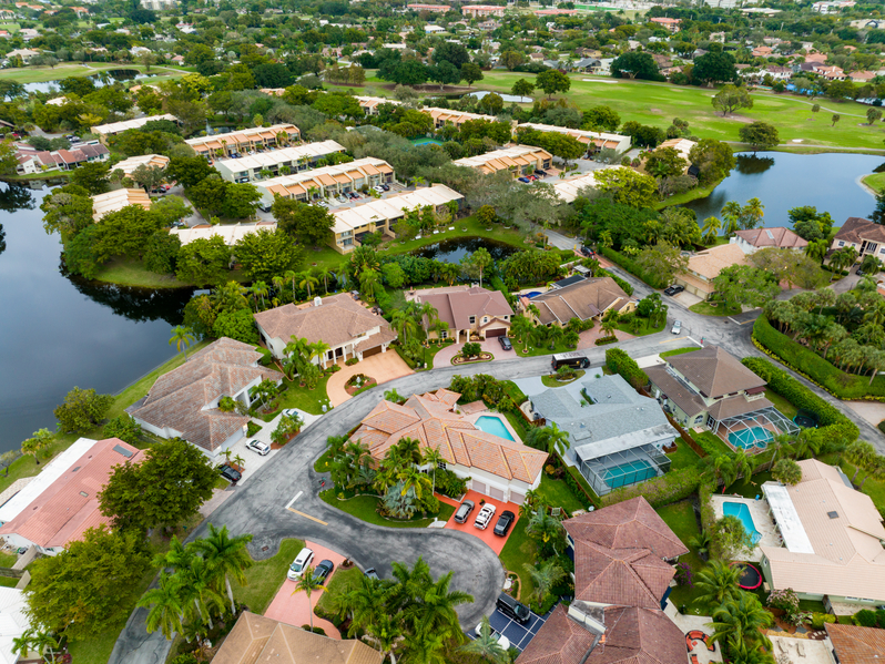 Aerial view of a suburban neighborhood with houses, swimming pools, green lawns, trees, and a pond. The area is surrounded by greenery and includes winding roads and a golf course in the background.