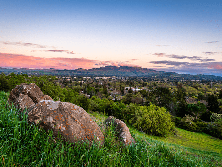 Large rocks sit in tall green grass overlooking a lush valley filled with trees and houses, with mountains in the background and a colorful sunset sky above.