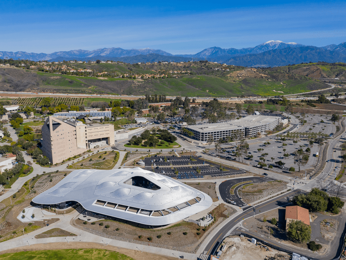 Aerial view of a futuristic, white, curved building surrounded by roads, parking lots, and greenery, with mountains in the background under a clear blue sky.