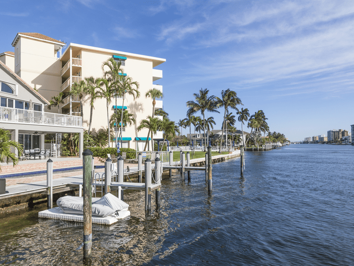 Waterfront view featuring a dock with boats, palm trees, and multi-story residential buildings along a calm canal under a clear blue sky.